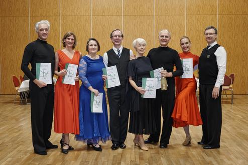 Gruppenfoto von Tänzern in festlicher Kleidung, die Urkunden in der Hand halten, in einem Saal mit Holzboden.
