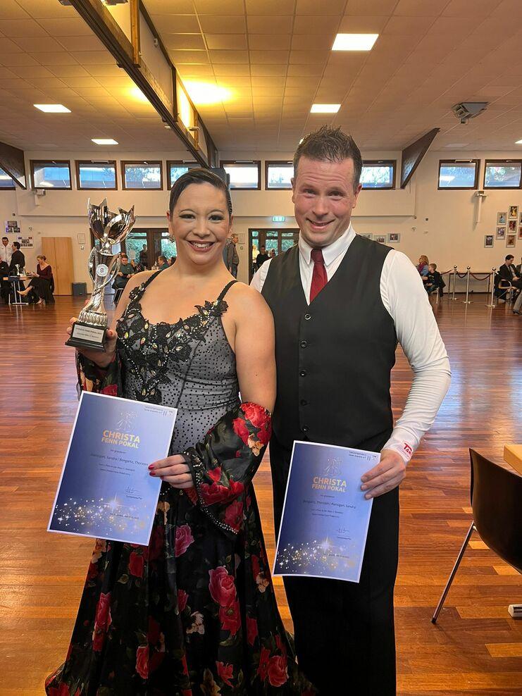 Tanzpaar hält einen Pokal und Urkunden in der Hand, lächelt in einem Veranstaltungssaal mit Holzboden.