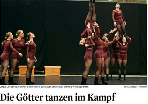 Acht Tänzerinnen in burgunderfarbenen Kostümen führen akrobatische Figuren auf einer Bühne vor schwarzem Hintergrund aus.