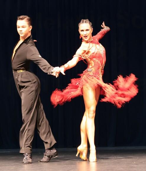 Tanzpaar in eleganter Pose, mit funkelnden Kostümen, auf einer Bühne vor schwarzem Hintergrund.