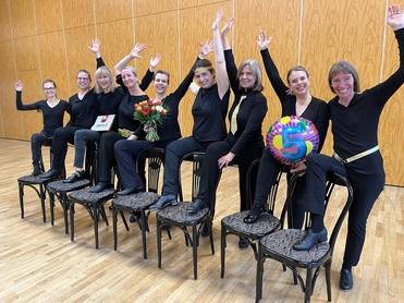 Acht Frauen in schwarzer Kleidung sitzen auf Stühlen, heben die Hände und feiern mit Blumen und einer „5“-Ballon.