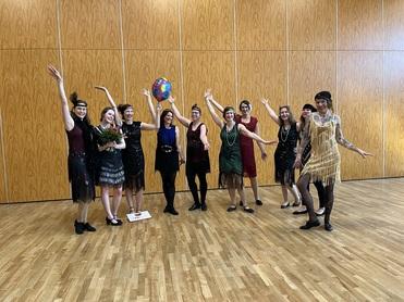 Gruppe von Frauen in glänzenden, fransigen Kleidern feiert in einem Raum, eine hält Blumen, eine andere einen Luftballon.