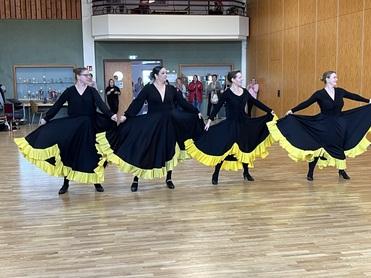 Vier Tänzerinnen in schwarzen Kleidern mit gelben Rüschen führen eine einstudierte Choreografie auf einem Parkettboden vor.
