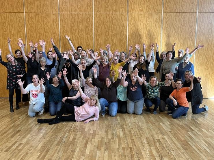 Gruppenfoto von 36 Frauen, die fröhlich mit erhobenen Händen stehen und sitzen, vor einer Holzpaneelwand.
