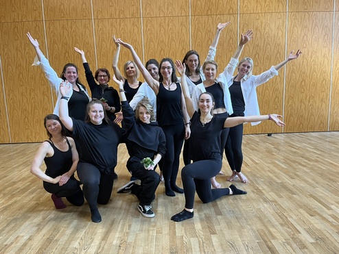 Gruppe von Frauen in Schwarz und Weiß posiert fröhlich in einem Tanzraum mit Holzfußboden.