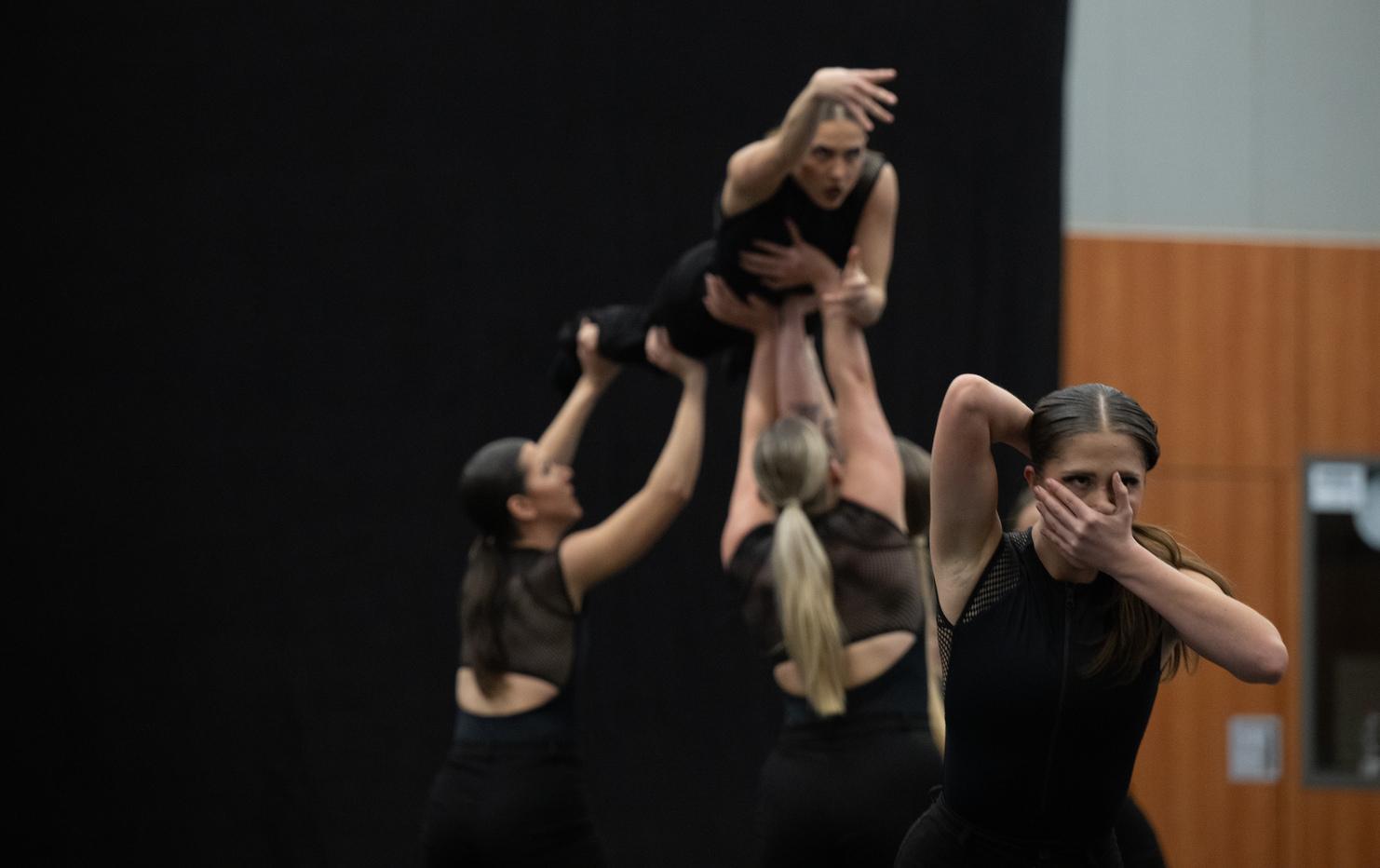 Sechs Tänzerinnen in Schwarz führen eine Hebefigur auf, während eine Tänzerin mit einem Handzeichen stoppt.
