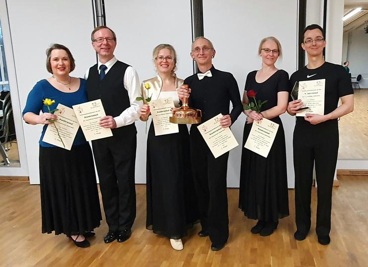 Sechsmal tanzende Paare präsentieren Urkunden und Blumen in festlicher Kleidung, stehend in einem Tanzsaal.