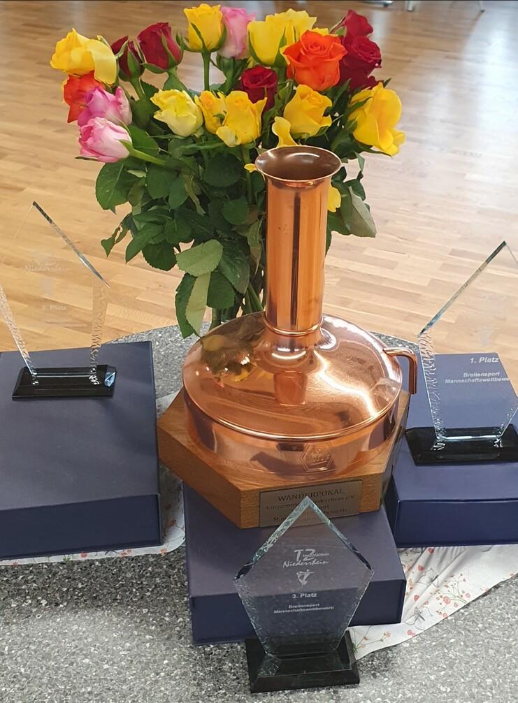 Kupferner Wanderpokal mit Blumen und drei Pokalen für den ersten bis dritten Platz auf einem Tisch.