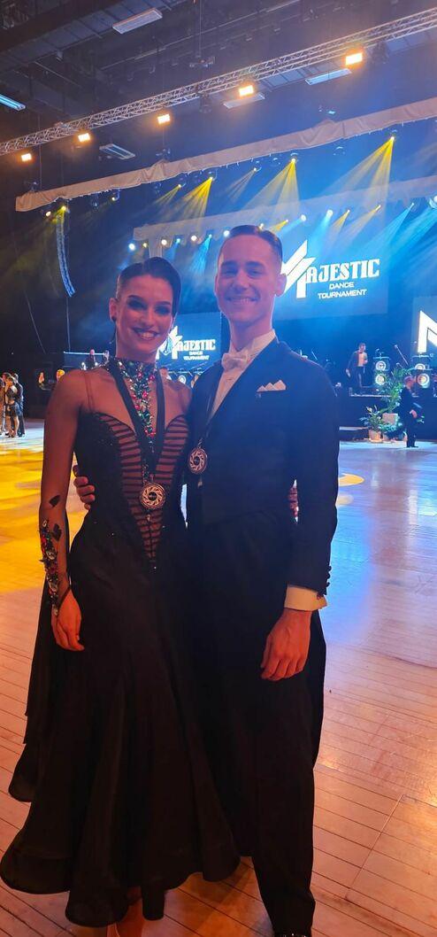 Tanzpaar in eleganter Tanzkleidung steht glücklich zusammen, Medaillen um den Hals, vor einer beleuchteten Bühne.