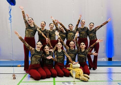 Gruppe von Tänzerinnen in bunter Kleidung posiert fröhlich vor einer Wand, mit einem Plüschgiraffen und einer Trophäe.