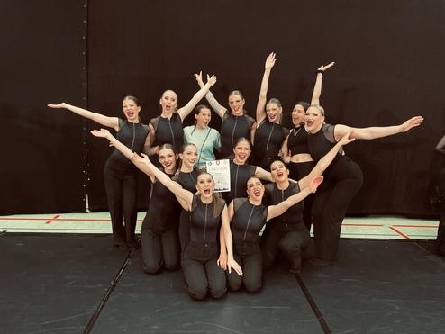 Gruppe von Tänzerinnen in schwarzer Kleidung, die freudig um ein Zertifikat posieren. Schwarzer Hintergrund.