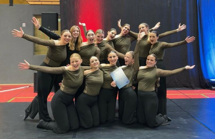 Gruppe von Tänzerinnen in olivefarbenen Outfits posiert fröhlich mit Urkunde auf einer Bühne.