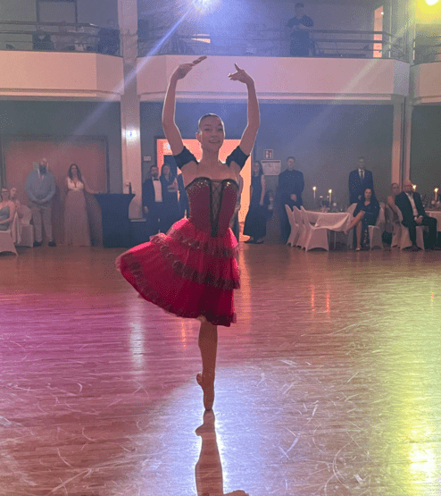 Tänzerin in einem roten Kleid zeigt eine elegante Pose auf einer Bühne, umgeben von applaudierendem Publikum.