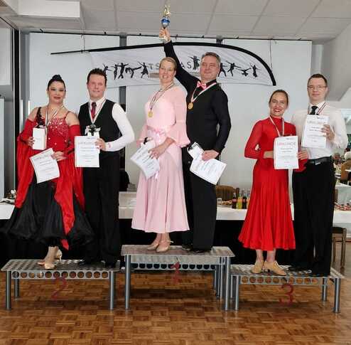 Fünf Tänzer stehen auf Treppen, halten Urkunden und Pokale; zwei tragen elegante Tanzkleider in Schwarz, Rot und Rosa.
