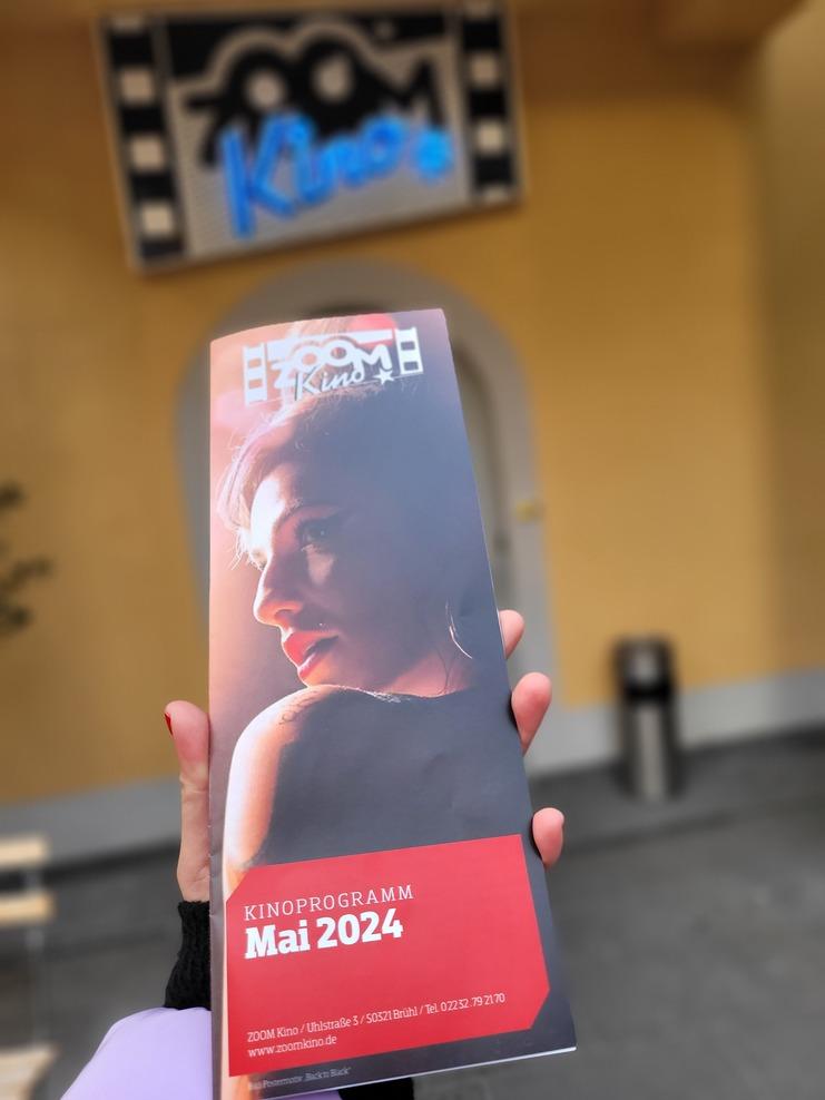 Hand hält ein Kinoprogramm für Mai 2024 mit Titel und Bild einer Frau, im Hintergrund das ZOOM Kino-Schild.