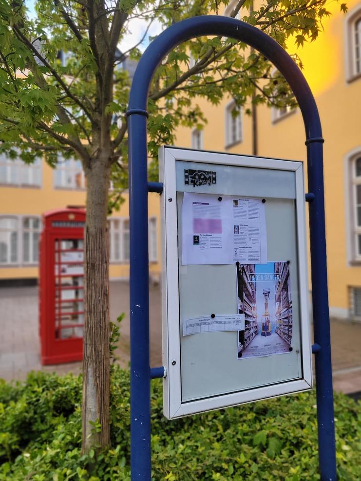 Informationsschild mit Ankündigungen, Plakaten und einem roten Telefonzelle im Hintergrund vor einem Gebäude.