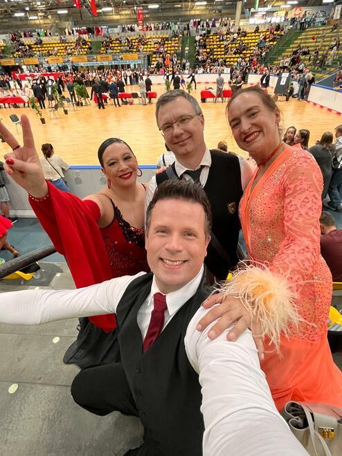 Vier Personen posieren in festlicher Tanzkleidung im Vordergrund, mit einer Tanzveranstaltung im Hintergrund.