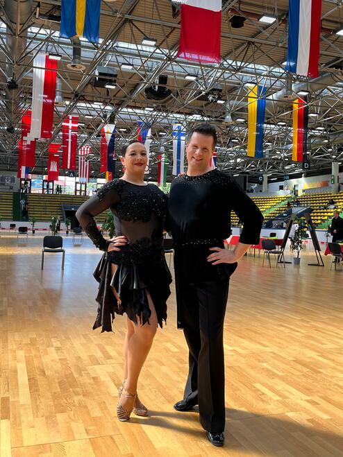 Tanzpaar in glitzernden Kostümen posiert in einer Turnhalle mit Fahnen an der Decke und Parkettboden.