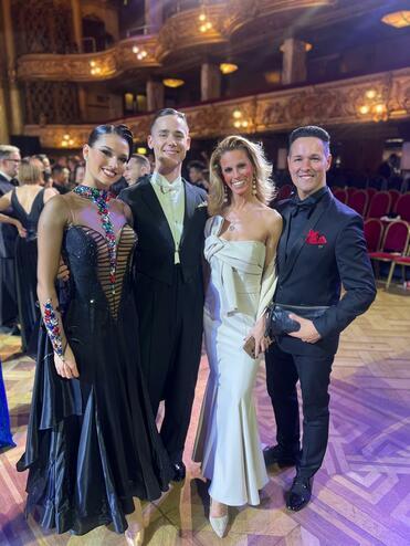 Vier elegant gekleidete Personen posieren lächelnd vor einem festlichen Hintergrund mit Goldverzierungen und Holzfußboden.