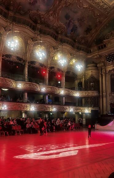 Elegant gestalteter Saal mit beleuchtetem Parkett, Zuschauer in roten Sesseln und einem Kind in Schwarz im Vordergrund.