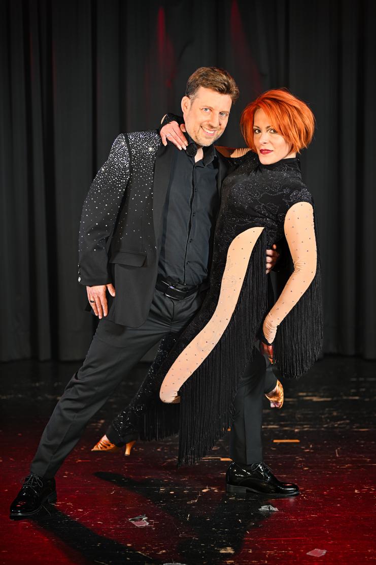 Tanzpaar in eleganter Kleidung, lächelt und posiert vor einem schwarzen Vorhang mit rotem Licht.