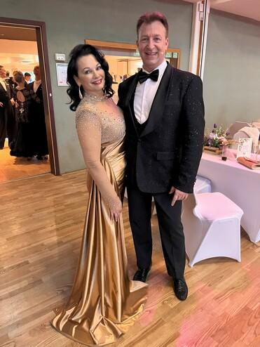 Paar in eleganter Abendgarderobe, sie trägt ein goldfarbenes Kleid, er einen glitzernden schwarzen Anzug.