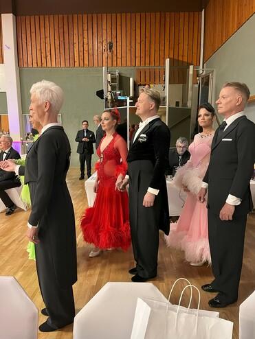 Fünf Tänzer in eleganter Kleidung stehen nebeneinander auf einer Tanzfläche, bereit für einen Wettbewerb.