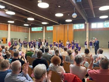 Tanzaufführung in einer Halle mit Zuschauern, Kindern in lila T-Shirts zeigen eine Choreografie. Applaus vom Publikum.
