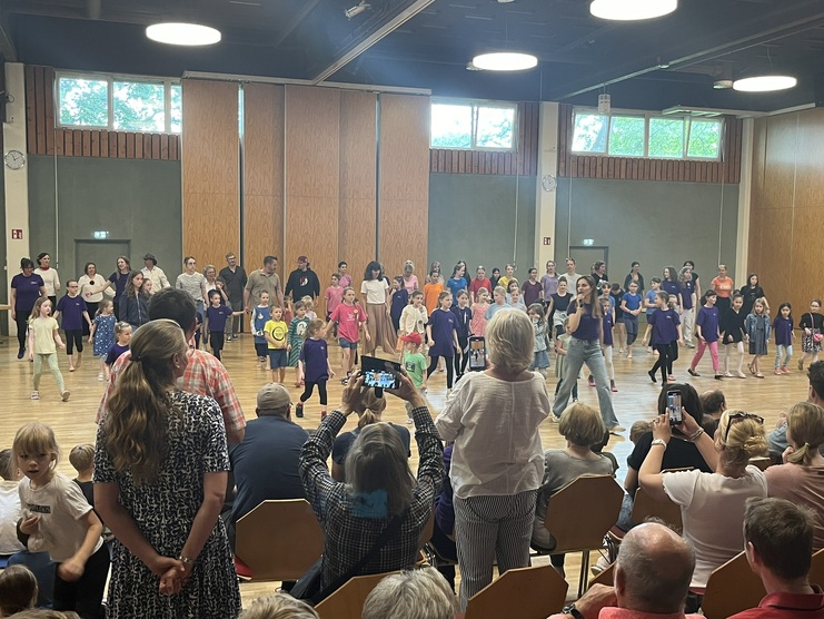 Gruppentanzauff&uuml;hrung in einer Halle mit Kindern und Erwachsenen in bunten Outfits, Zuschauer beobachten gespannt.
