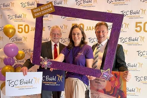 Drei Personen posieren mit einer glitzernden Rahmenattrappe vor einer Wand mit „50 Jahre TSC Brühl“-Dekoration und Luftballons.