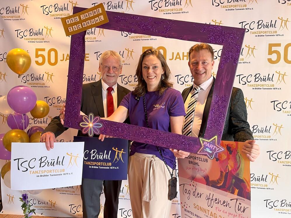 Drei Personen posieren mit einer glitzernden Rahmenattrappe vor einer Wand mit &bdquo;50 Jahre TSC Br&uuml;hl&ldquo;-Dekoration und Luftballons.