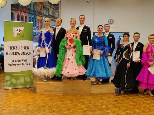 Gruppenshot von zehn Tänzern in festlicher Kleidung, auf einem Podium stehend, mit einer Glückwunschtafel im Hintergrund.