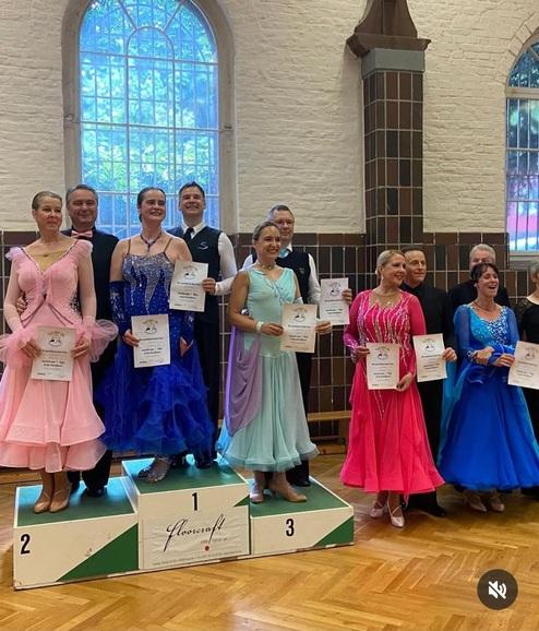 Gruppe von tanzenden Paaren in eleganter Kleidung mit Urkunden auf dem Siegerpodest in einem Innenraum.