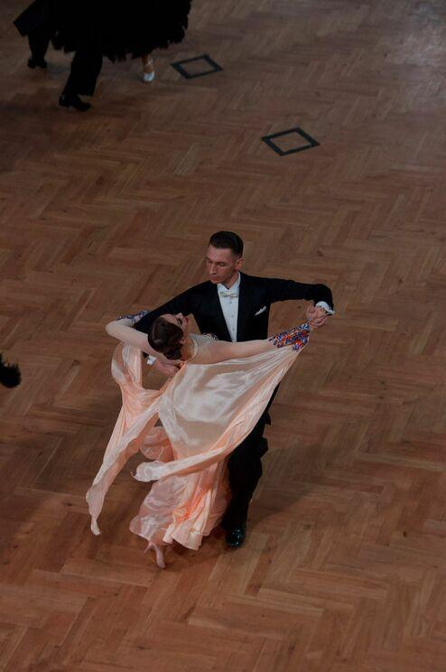 Tanzpaar bei einem Wettbewerb, die Frau in einem fließenden, hellen Kleid, der Mann im klassischen Anzug auf Holzboden.