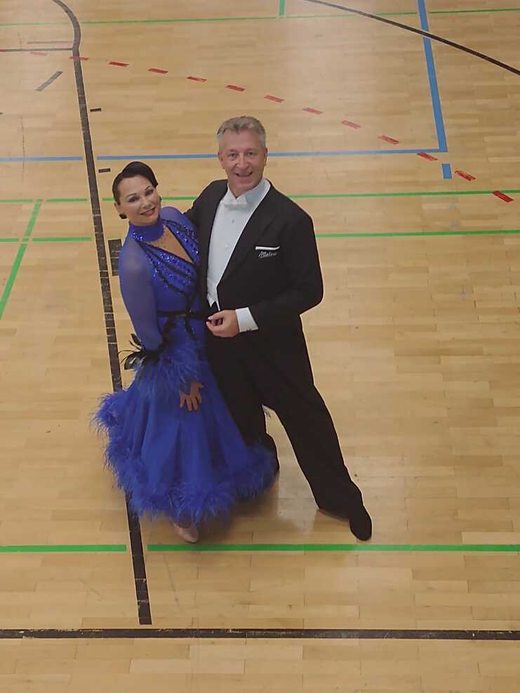 Tanzpaar in eleganter Kleidung posiert auf einer Sportfläche mit Holzfußboden, umgeben von farbigen Linien.