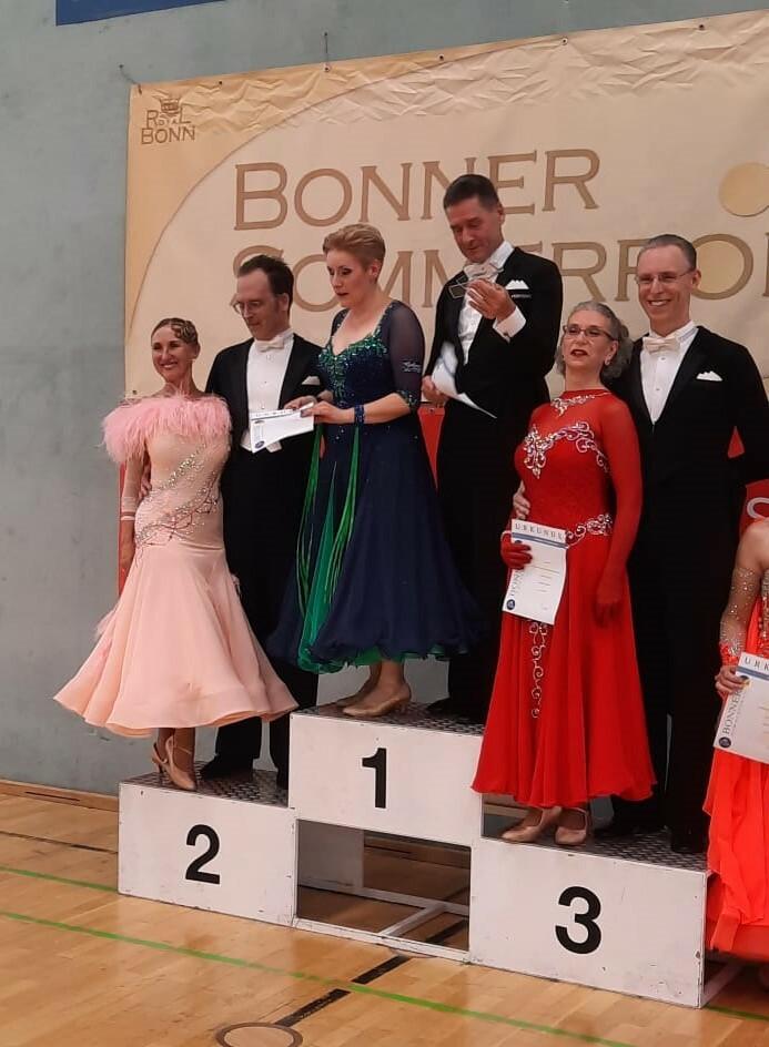 Sechs Tänzer in eleganten Kleidern stehen auf dem Podium, während eine Urkunde überreicht wird. Hintergrund mit Banner.