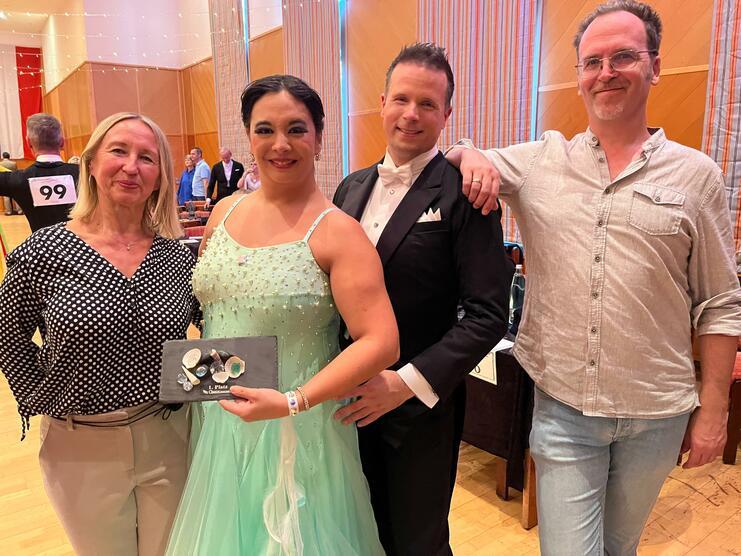 Vier Personen posieren in formeller Kleidung bei einer Tanzveranstaltung, eine Frau hält eine dekorierte Plakette in der Hand.