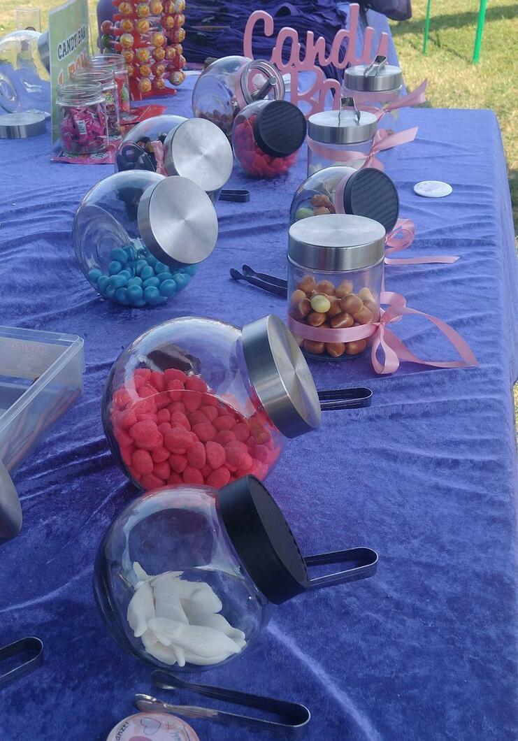 Bunte Bonbongläser mit verschiedenen Süßigkeiten auf einem lila Tischdecken, mit Löffeln zur Entnahme.