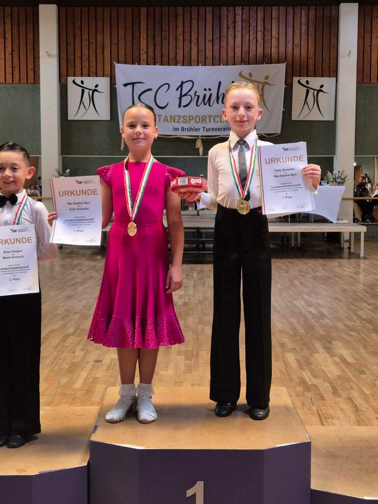 Drei Kinder stehen auf dem Siegertreppchen mit Medaillen und Urkunden. Die erste Platzierte trägt ein rosa Kleid.