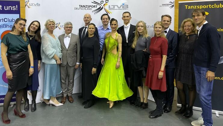 Gruppenfoto von Tanzsportfans vor einem Banner, eine Tänzerin in leuchtend grünem Kleid steht im Vordergrund.