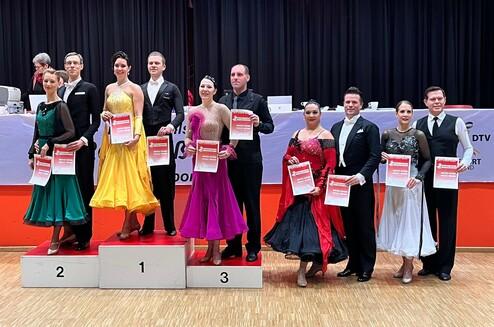 Tanzpaare auf dem Siegertreppchen halten Urkunden in der Hand, dekoriert mit bunten Kleidern und Anzügen in einem Wettbewerbsraum.