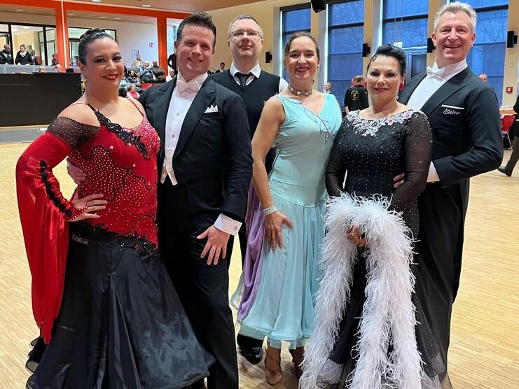 Sechs Tanzpaare in eleganter Kleidung posieren in einem Tanzsaal, umgeben von Holzfußboden und Zuschauern im Hintergrund.