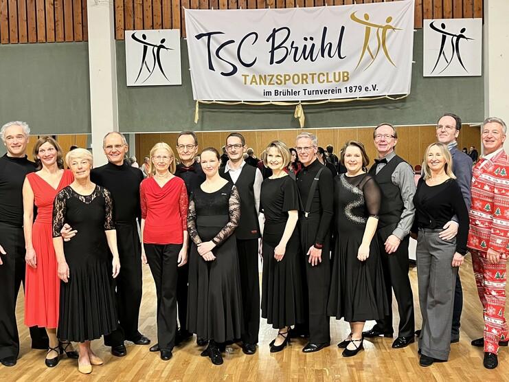Gruppe von Tänzern in festlicher Kleidung posiert vor Banner des TSC Brühl in einer Tanzhalle.
