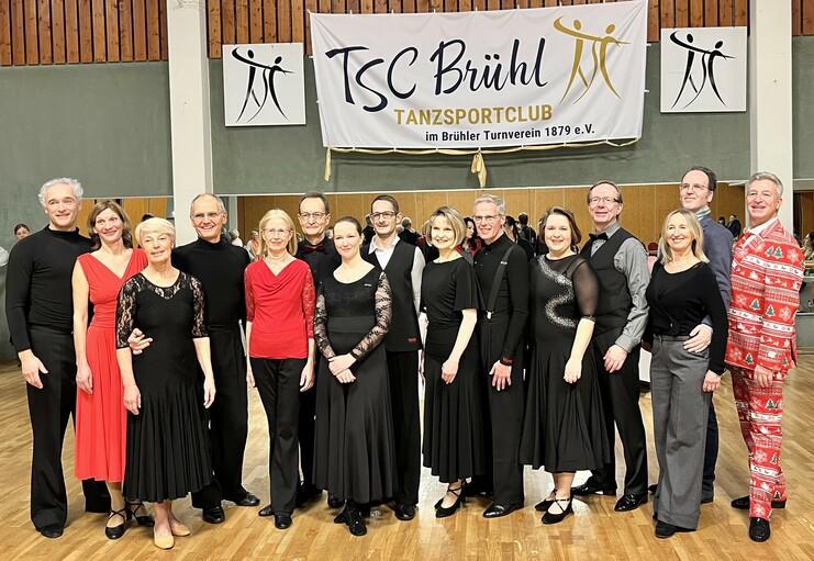 Gruppe von Tänzern in festlicher Kleidung posiert vor Banner des TSC Brühl in einer Tanzhalle.