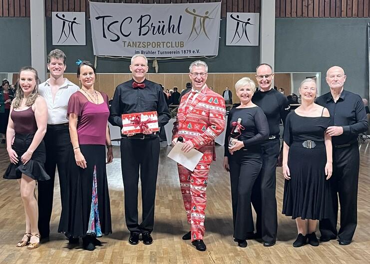 Gruppe von Tänzern in festlicher Kleidung pose vor Banner des Tanzsportclubs Brühl, einige halten Geschenke und Pokale.