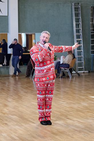 Mann in festlichem rot-grünem Outfit singt mit Mikrofon in einem Saal mit Sitzplätzen im Hintergrund.