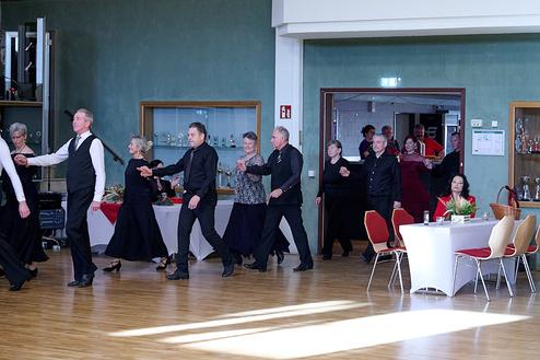 Tanzende Personen in eleganter Kleidung bewegen sich in einem festlichen Saal mit dekorierten Tischen im Hintergrund.