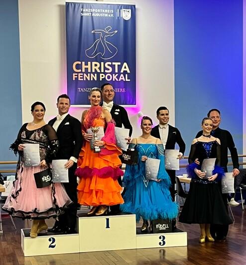 Sieben Tänzerinnen und Tänzer stehen auf dem Siegertreppchen, gehaltenen Pokalen und Urkunden in einem Tanzwettbewerb.