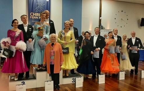 Sieger und Teilnehmer eines Tanzturniers stehen auf Podesten in festlicher Kleidung, umrahmt von Publikum und Auszeichnungen.