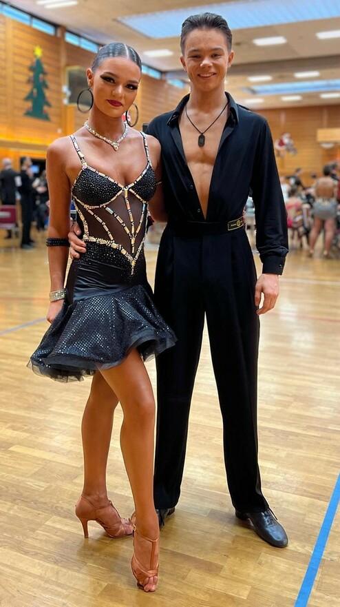 Tanzpaar in eleganten Outfits, die auf einer Tanzfläche posieren, umgeben von einem Wettbewerbsumfeld.